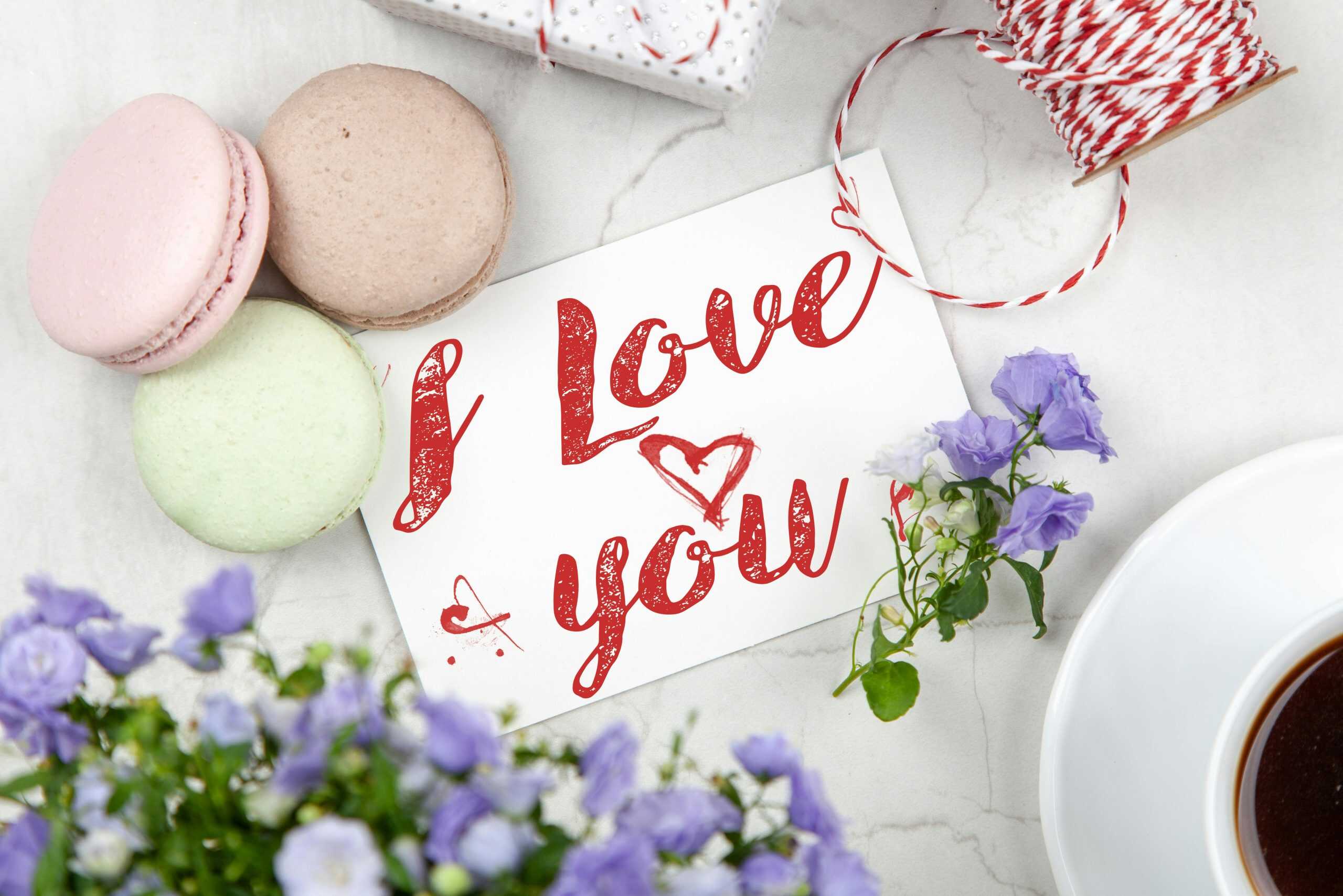 An artistic flat lay of an 'I Love You' card surrounded by macarons, flowers, and coffee, perfect for Valentine's Day.